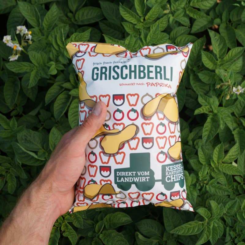 Grischberli Paprika Chips 120g package held in hand with green foliage background.